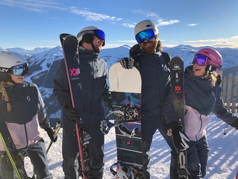 Vier Personen mit Ski und Snowboard am Pistenrand, dahinter das Tal