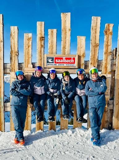 Sechs Personen in Skikleidung vor einem Holzzaun mit dem Ortsschild Saalbach