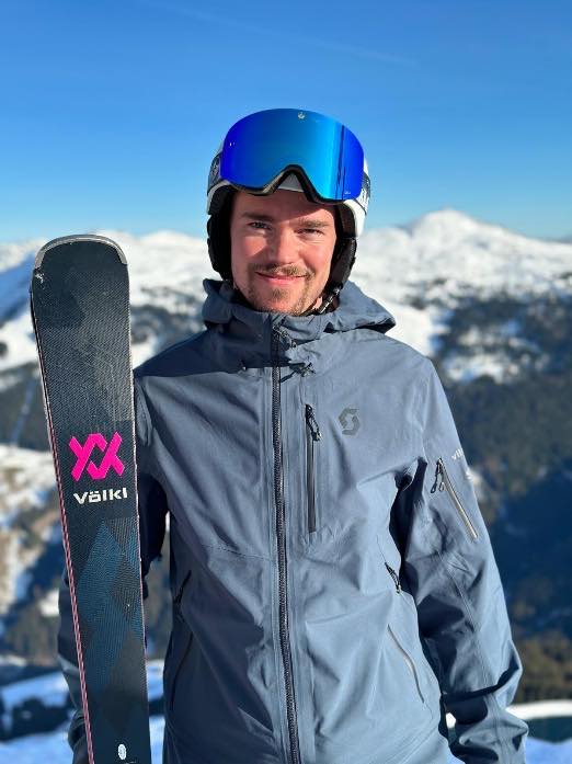 Person mit Helm und Skibrille, die Ski geschultert, dahinter das Tal