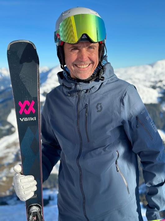 Person mit Helm und Skibrille, die Ski geschultert, dahinter das Skigebiet