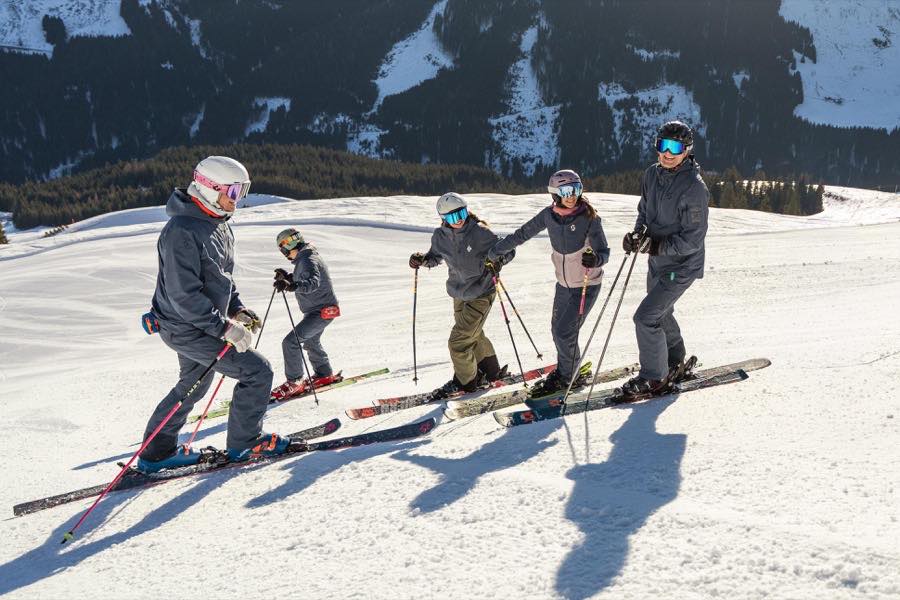 Fünf Personen mit Ski nebeneinander am Hang, dahinter bewaldete Berge